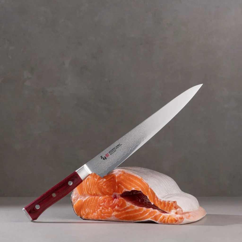 En kniv fra Mcusta Zanmai Classic Pro Damascus-serien står i et laksestykke, mot grå bakgrunn.
