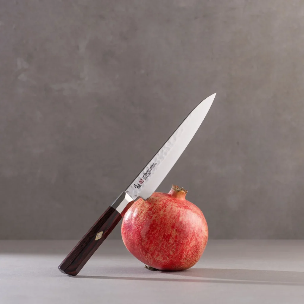En kniv med hamret blad fra Mcusta Zanmai Supreme Hammered-serien står i et granateple, mot grå bakgrunn.